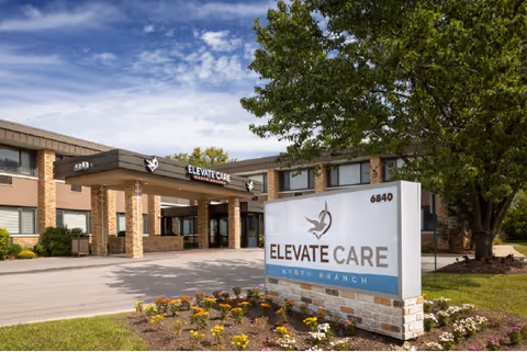 Exterior view of Elevate Care North Branch facility showing the entrance with a covered drop-off area, a large sign with the facility name and logo, a two-story brick building, a tree, and a flower bed in the foreground under a partly cloudy sky.