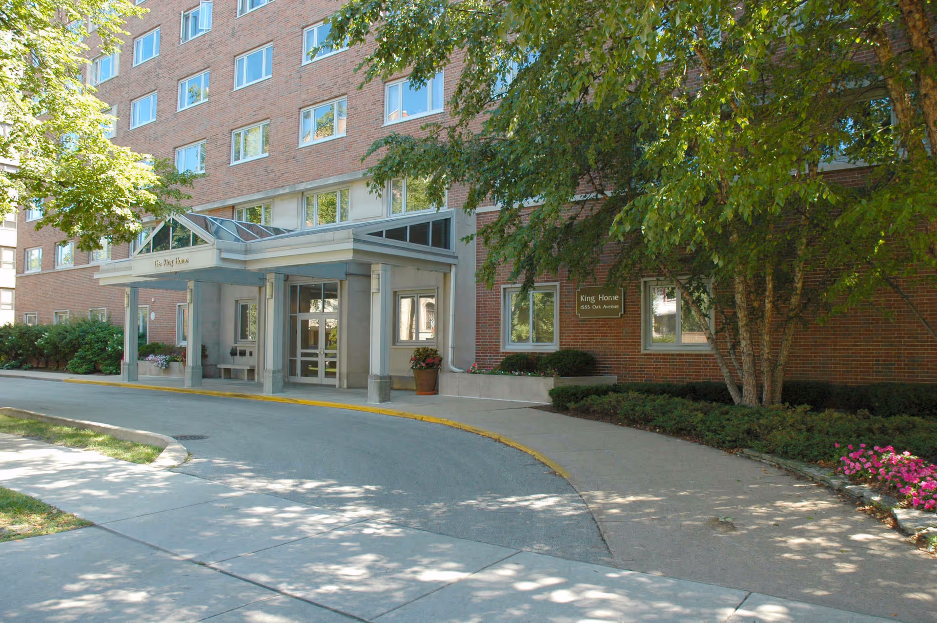 Exterior view of a multi-story brick building with a covered entrance labeled 'The King Home'. There are trees and shrubs around the entrance, with a curved driveway and sidewalk in front.