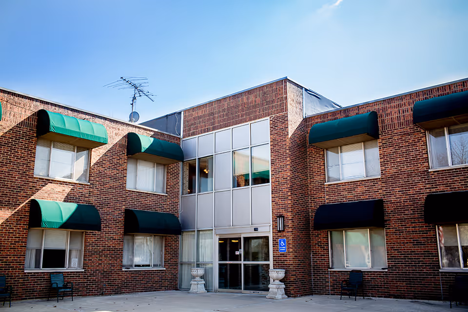 Exterior view of a two-story brick building with green and black awnings over the windows. The entrance features glass doors and a handicap accessible sign is visible near the door. The sky is clear and blue above the building.