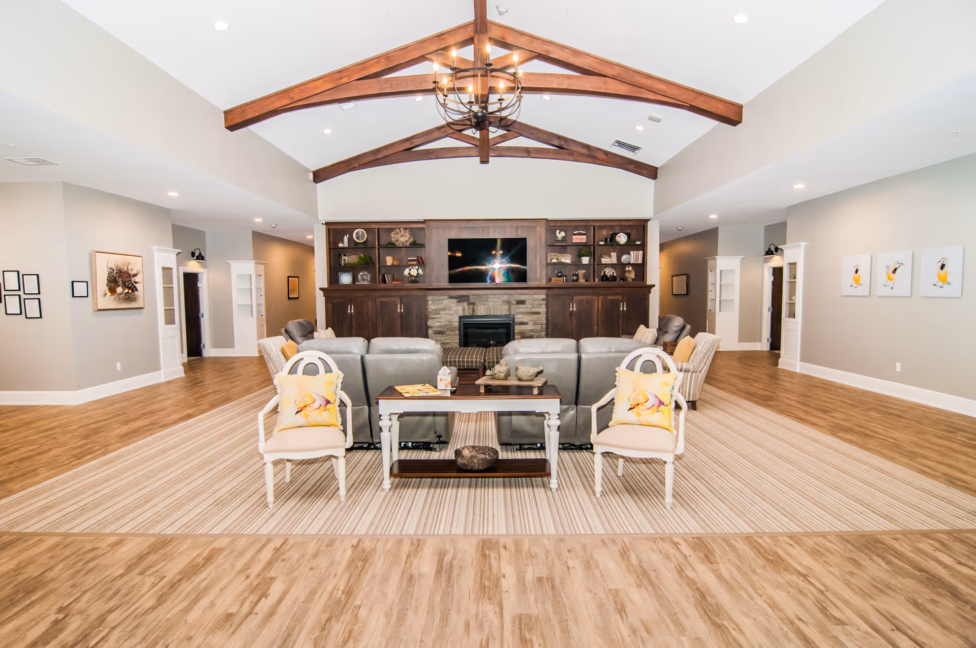 A spacious senior living facility common area with wooden flooring and a large striped rug. The room features a seating arrangement with two white chairs with yellow floral cushions, a wooden coffee table, and several gray sofas facing a built-in wooden entertainment center with a TV and decorative shelves. The ceiling has exposed wooden beams and a chandelier, and the walls are decorated with framed artwork.