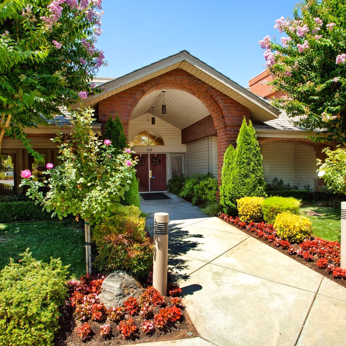 Front entrance of a building with a covered porch supported by brick columns, surrounded by well-maintained landscaping including flowering bushes, green shrubs, and trees under a clear blue sky.
