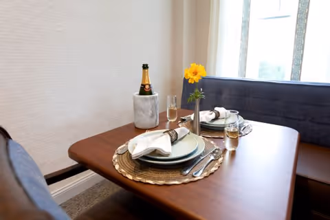 A small dining table set for two with plates, napkins, silverware, glasses with a light beverage, a bottle of champagne in a marble cooler, and a single yellow flower in a vase. The table is next to a window with blue cushioned bench seating.