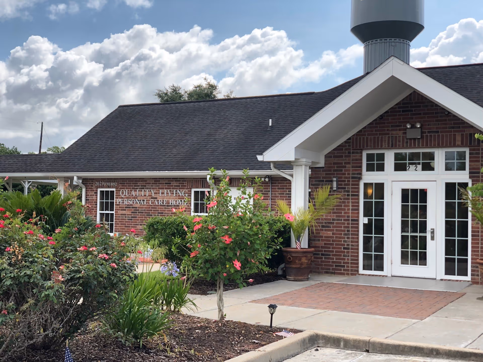 Exterior view of Quality Living Murphy, a brick building with a dark shingled roof, white framed double glass doors, and a small garden with flowering plants and shrubs in front. The sky is partly cloudy.