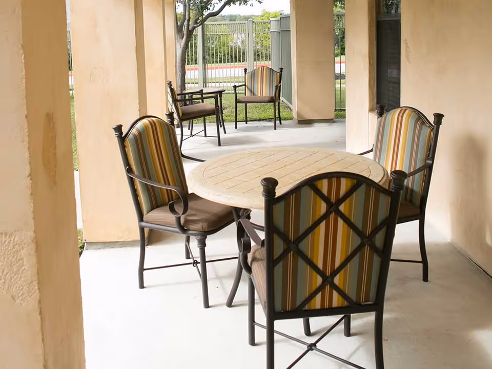 Outdoor covered patio area with a round table and four striped cushioned chairs, with two additional chairs and a small table in the background near a metal fence and greenery.