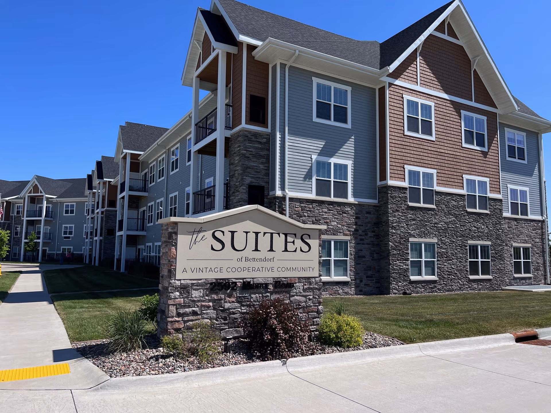 Exterior view of The Suites of Bettendorf, a multi-story residential building with stone and siding facade, multiple windows, and balconies under a clear blue sky. A stone sign in front reads 'The Suites of Bettendorf, A Vintage Cooperative Community.'