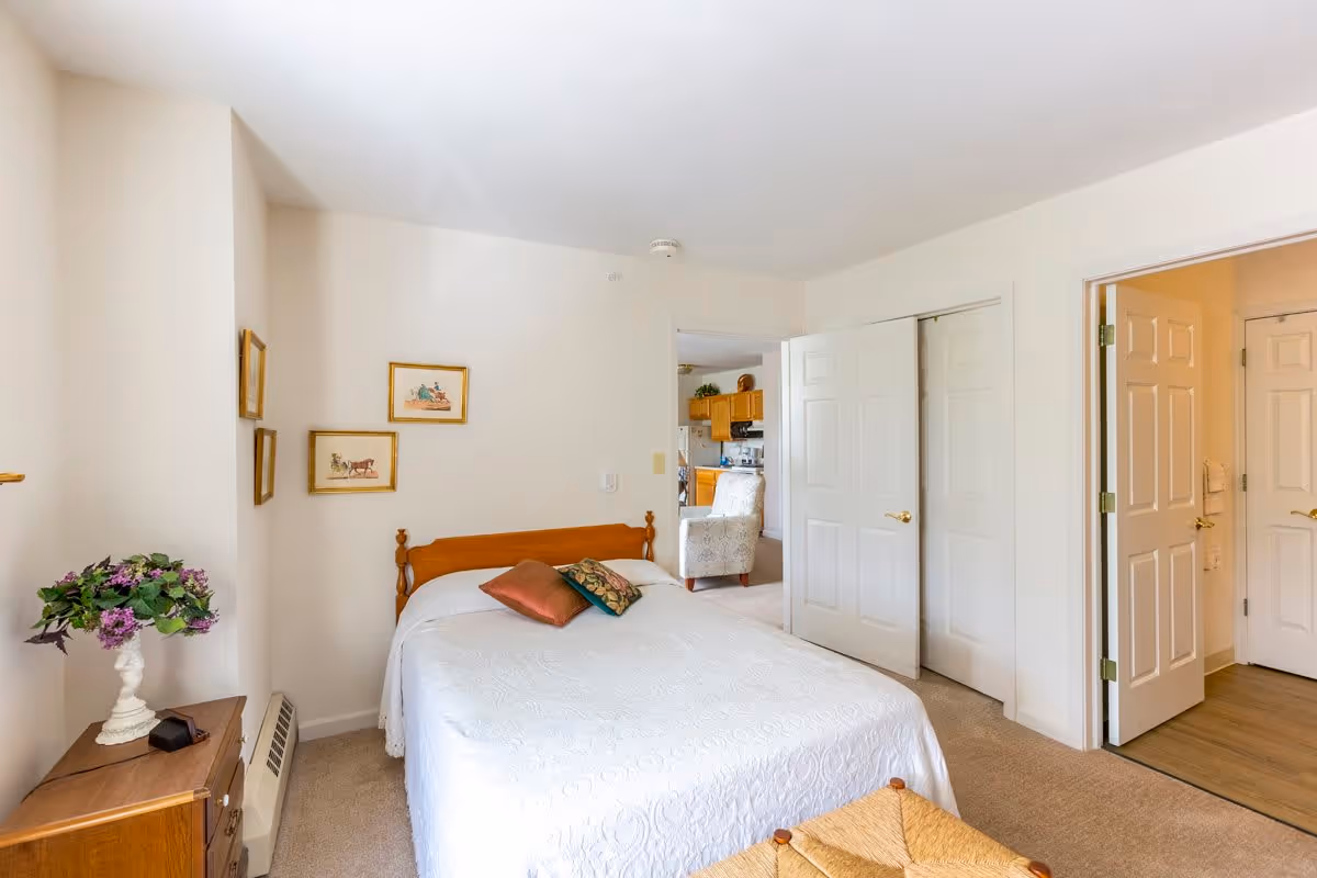 A bedroom with a wooden bed frame and white bedspread, two decorative pillows, a wooden nightstand with a vase of purple flowers, and three framed pictures on the wall. The room has beige carpet and white walls. Two open doors reveal a glimpse of a kitchen area and a hallway.