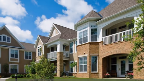 Exterior view of a senior living facility with brick and beige siding, multiple windows, balconies, and a well-maintained lawn under a partly cloudy blue sky.