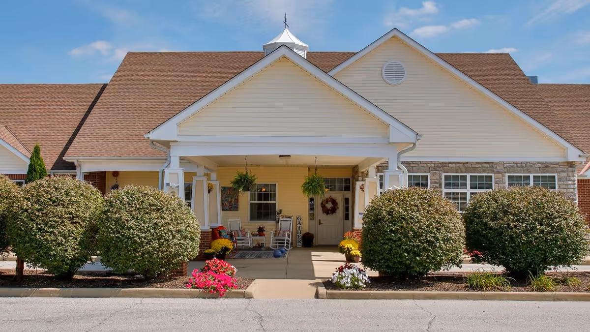 Front exterior view of Bickford of Lafayette facility featuring a covered entrance with white pillars, a peaked roof, hanging plants, rocking chairs, and neatly trimmed bushes along the walkway under a blue sky.