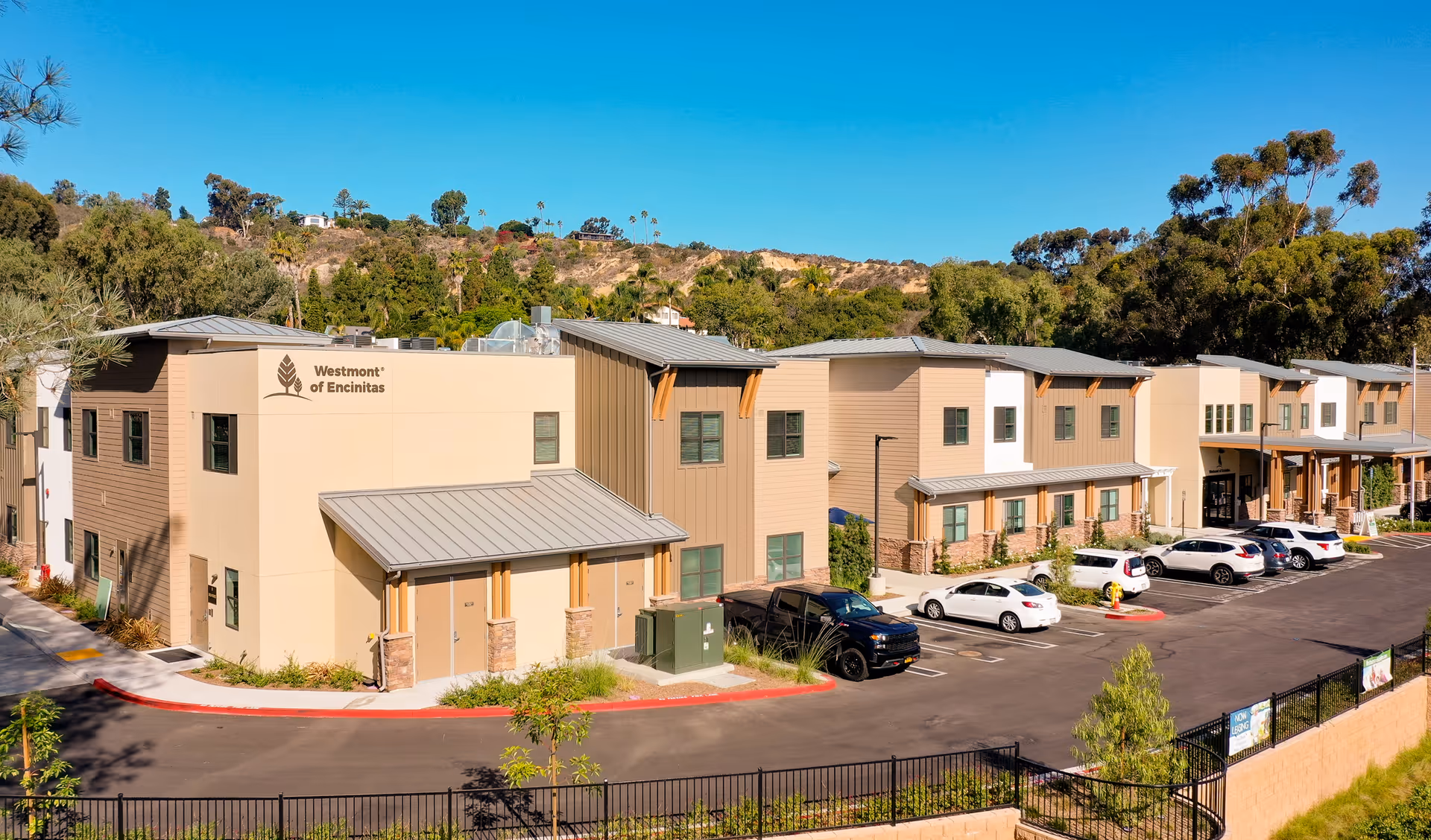 Exterior view of Westmont of Encinitas senior living facility showing a modern two-story building with beige and brown siding, multiple windows, a parking lot with several parked cars, and a backdrop of trees and hills under a clear blue sky.