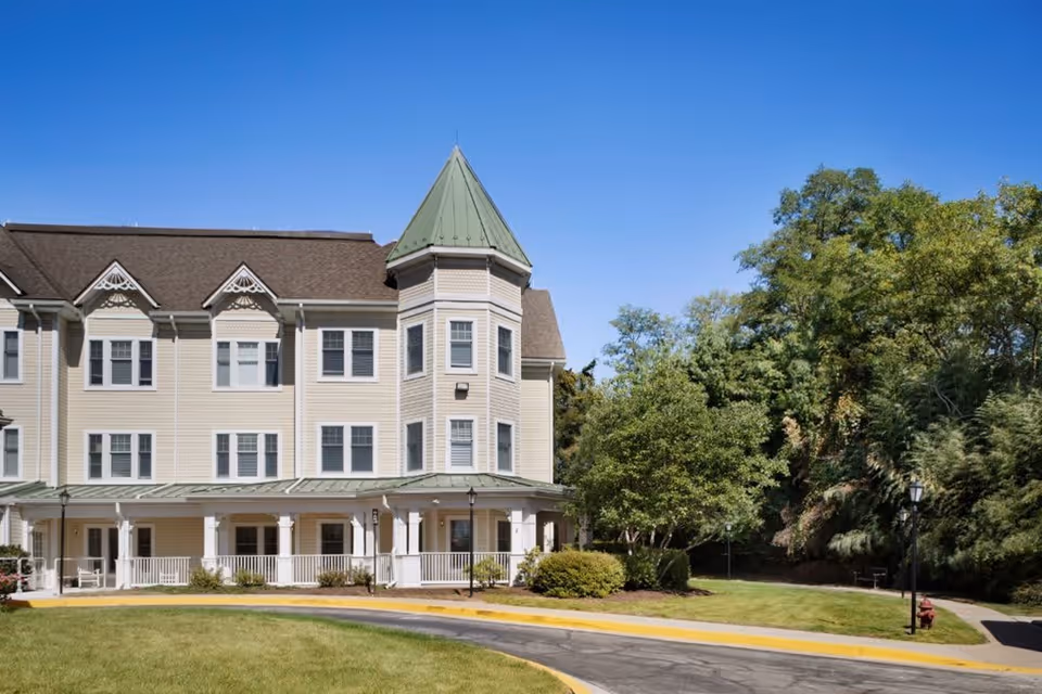 Exterior view of a multi-story senior living facility building with beige siding, a green pointed roof tower, multiple windows, and a covered porch area. The building is surrounded by a well-maintained lawn, trees, and a paved driveway under a clear blue sky.