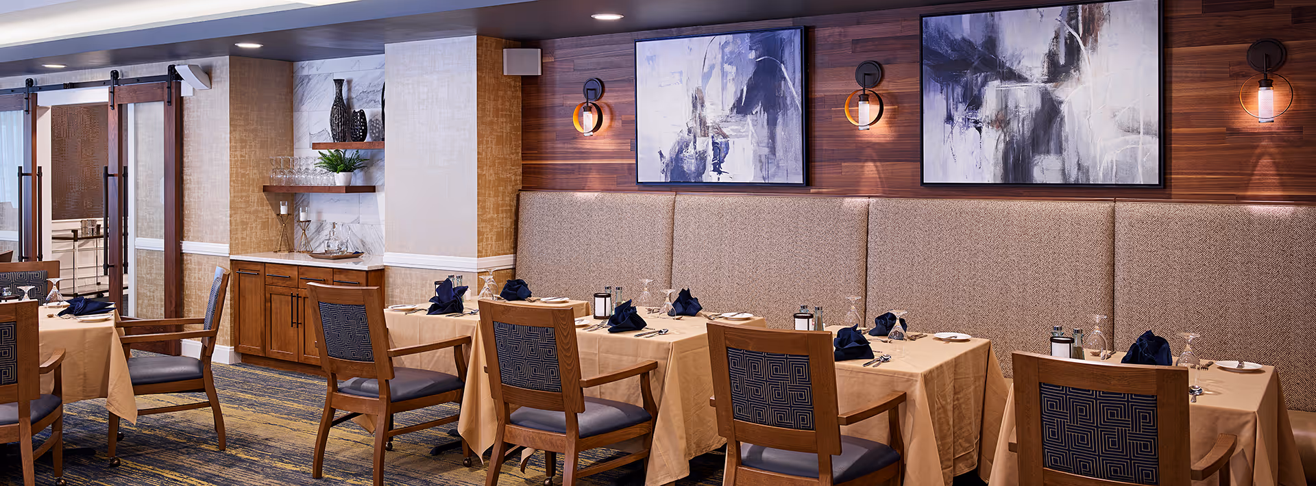 A dining room with tables covered in beige tablecloths, set with plates, silverware, glasses, and navy blue napkins. Wooden chairs with patterned blue cushions are arranged around the tables. The room features a wooden accent wall with two abstract paintings and wall sconces. There is a cabinet with shelves holding decorative vases and glassware, and sliding barn-style doors in the background.