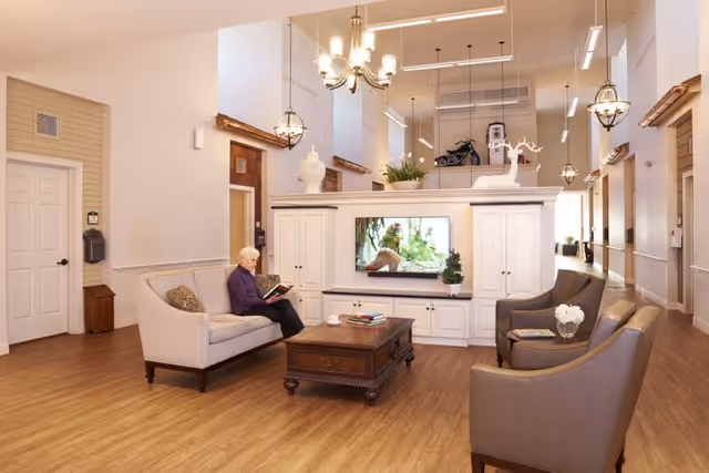 A bright and spacious common area in a senior living facility with wooden flooring, white walls, and high ceilings. There is a white entertainment center with a TV displaying a nature scene, flanked by decorative items including a white deer figurine and plants. Comfortable seating includes a beige sofa where an elderly woman is reading a book, and two brown armchairs with a small table holding a flower vase. The room is illuminated by hanging chandeliers and pendant lights, with a hallway visible in the background.