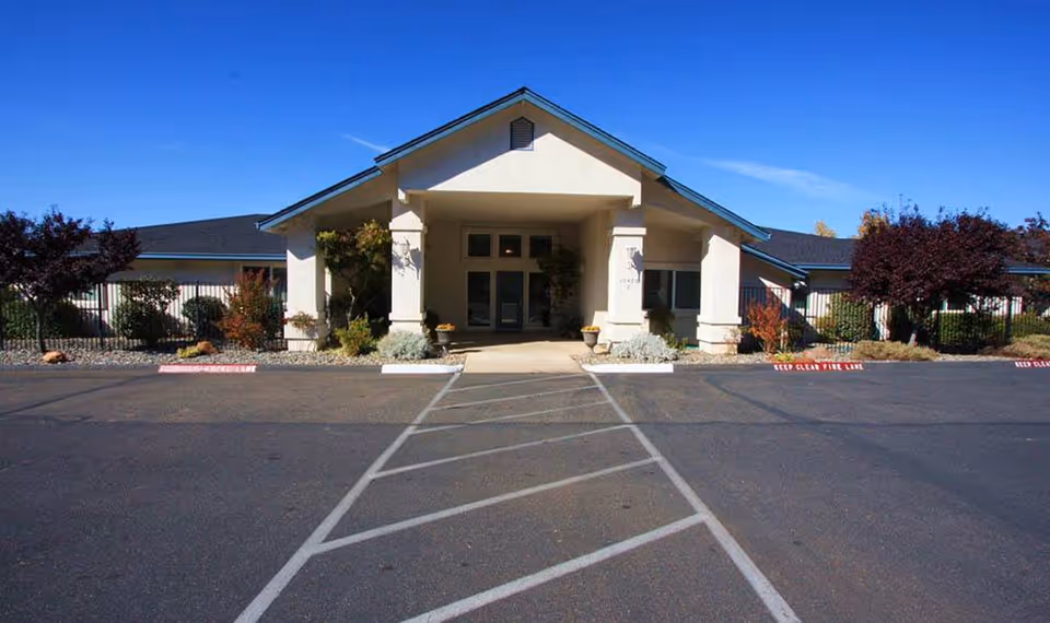 Front exterior view of Oak Terrace Memory Care facility with a covered entrance, surrounded by landscaped bushes and trees under a clear blue sky.