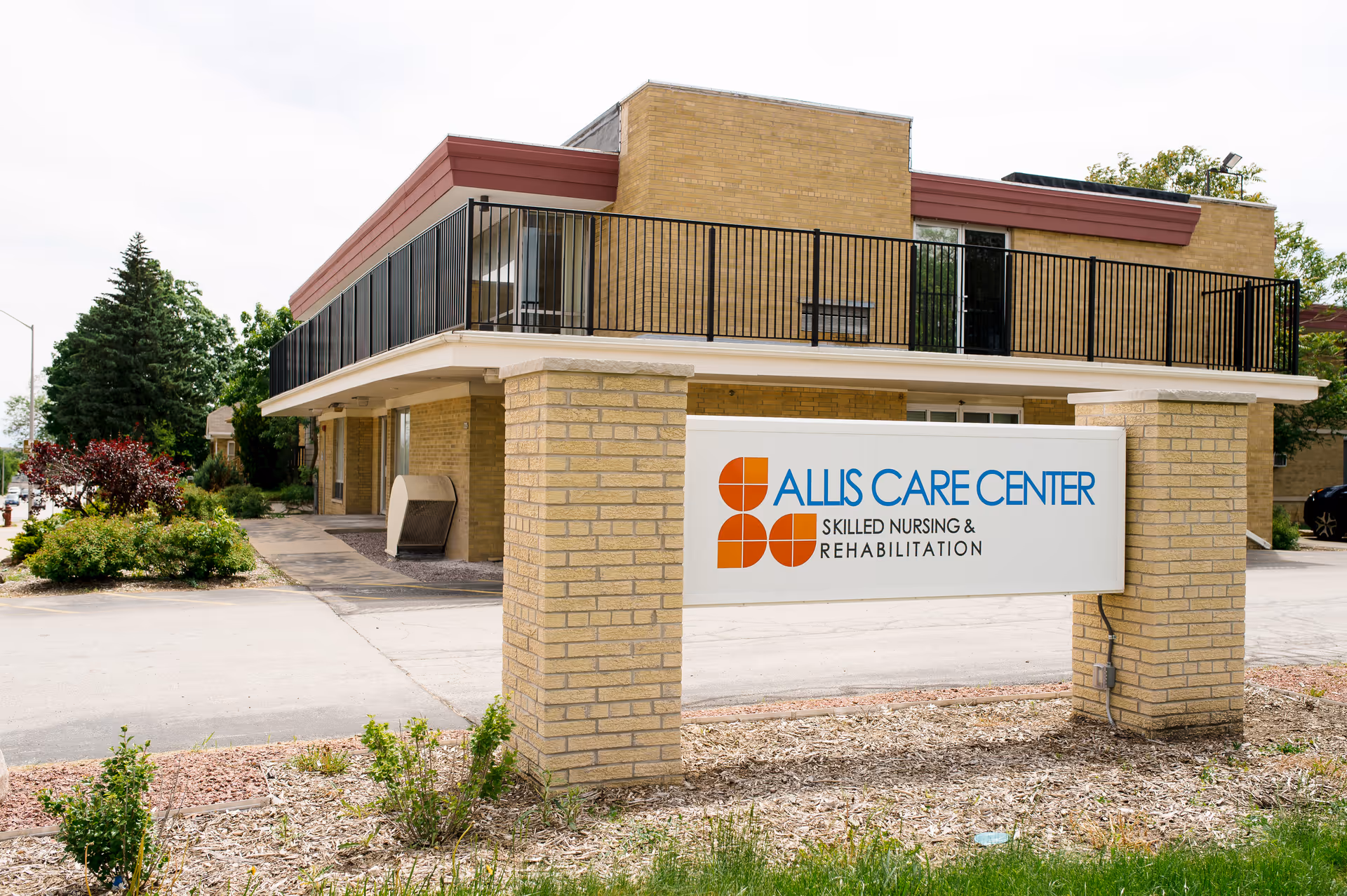 Exterior view of Allis Care Center, a skilled nursing and rehabilitation facility, showing a two-story brick building with a balcony and a large sign in front displaying the facility's name and logo.