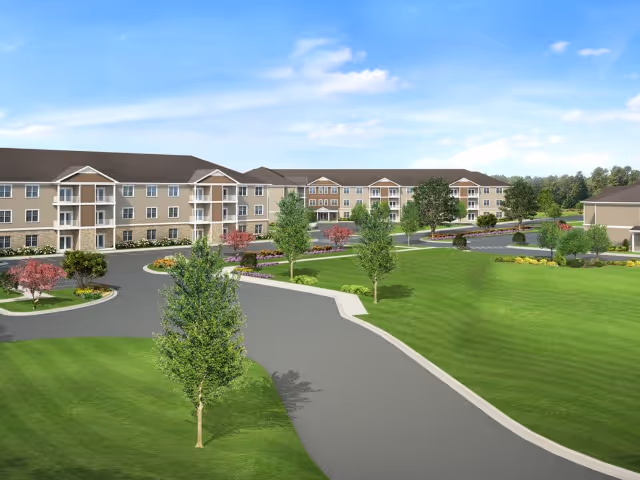 Exterior view of Carlton Hollow Senior Apartments showing a large, multi-story residential building with beige siding and stone accents. The building has multiple balconies and is surrounded by well-maintained green lawns, trees, and landscaped areas. A paved driveway curves through the front of the property under a partly cloudy blue sky.