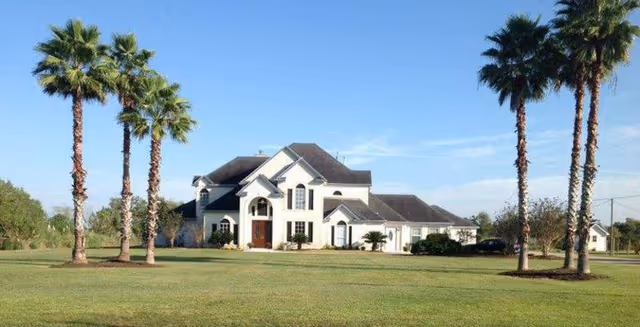 A large white two-story house with a dark roof situated on a spacious green lawn. There are several tall palm trees in the foreground and a clear blue sky above.