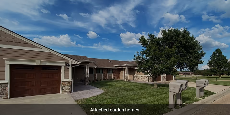 Attached garden homes with brown garage doors, a green lawn, trees, and a clear blue sky with some clouds.