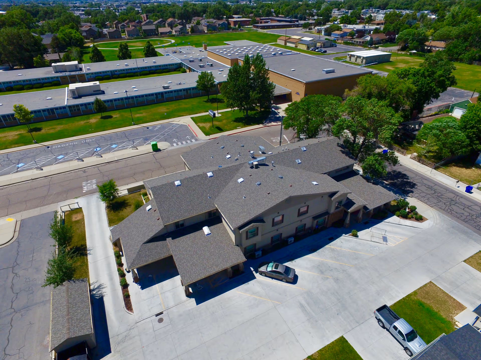 Aerial view of a senior living facility named Rosewood Assisted Care, showing a large building with a gray roof, surrounding parking lots with a few parked cars, nearby trees, and adjacent buildings in a suburban area.