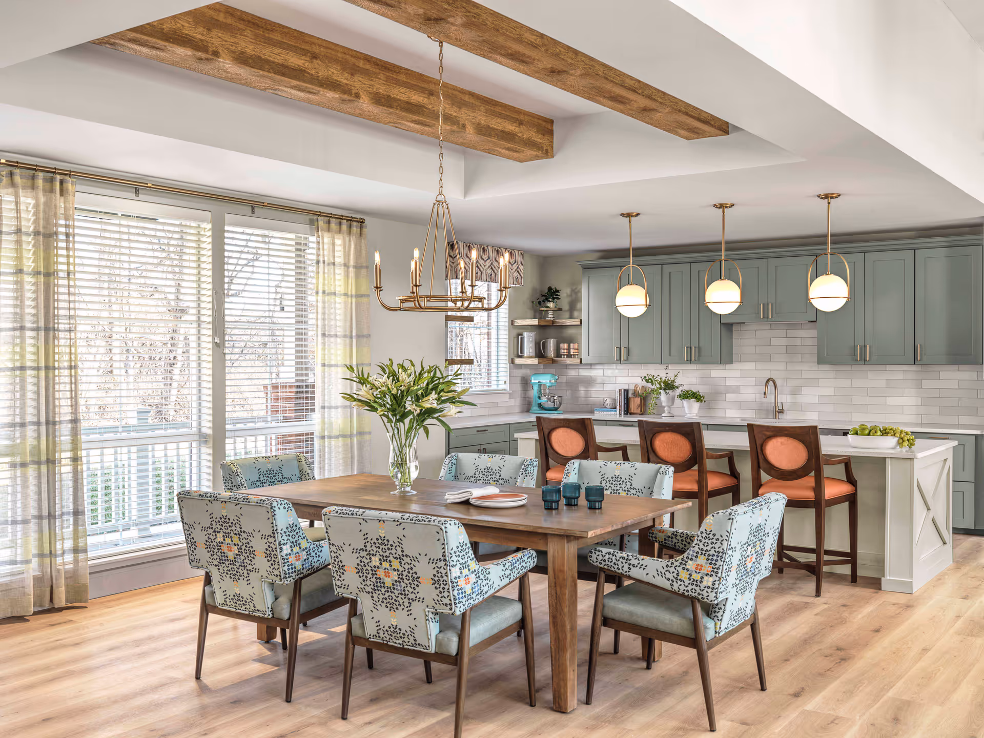 A bright and spacious dining area with a wooden table surrounded by six upholstered chairs with patterned fabric. Behind the dining table is a kitchen with green cabinetry, a white subway tile backsplash, and a white island with three bar stools featuring orange cushions. The room has large windows with sheer curtains, wooden ceiling beams, and modern pendant lighting above the island and a chandelier above the dining table.