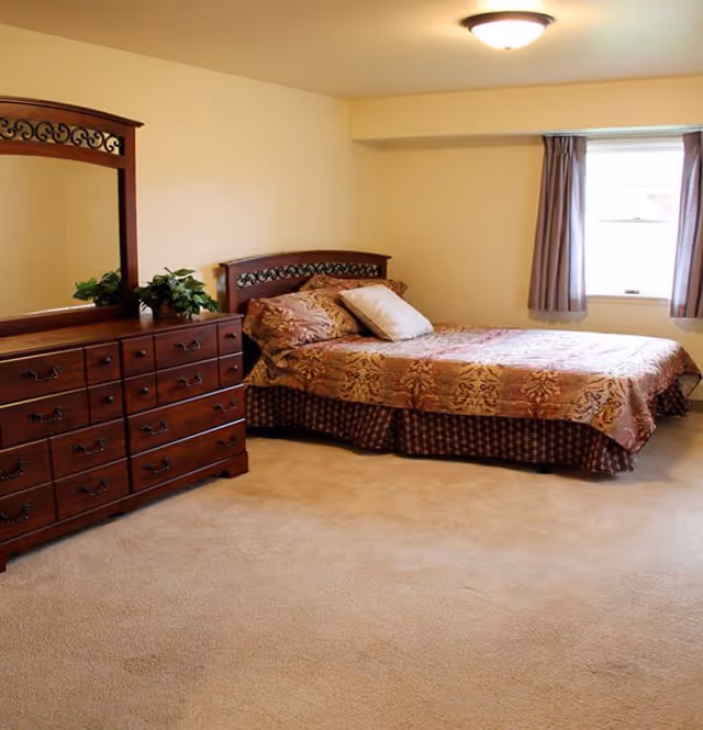 A bedroom with a double bed covered in a patterned bedspread and two pillows. There is a wooden dresser with a large mirror and decorative plants on top. A window with curtains allows natural light into the room, which has beige walls and carpeted flooring.