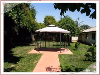 Outdoor garden area with a paved walkway leading to a gazebo with a white canopy. The area is surrounded by green grass, trees, and bushes, with a building visible on the right side.