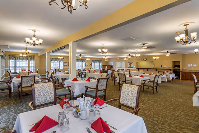 A spacious dining room with multiple tables covered in white tablecloths, each set with red folded napkins, glasses, and silverware. The room has patterned carpet, beige walls, several chandeliers, large windows with curtains, and wooden chairs with cushioned seats and backs.