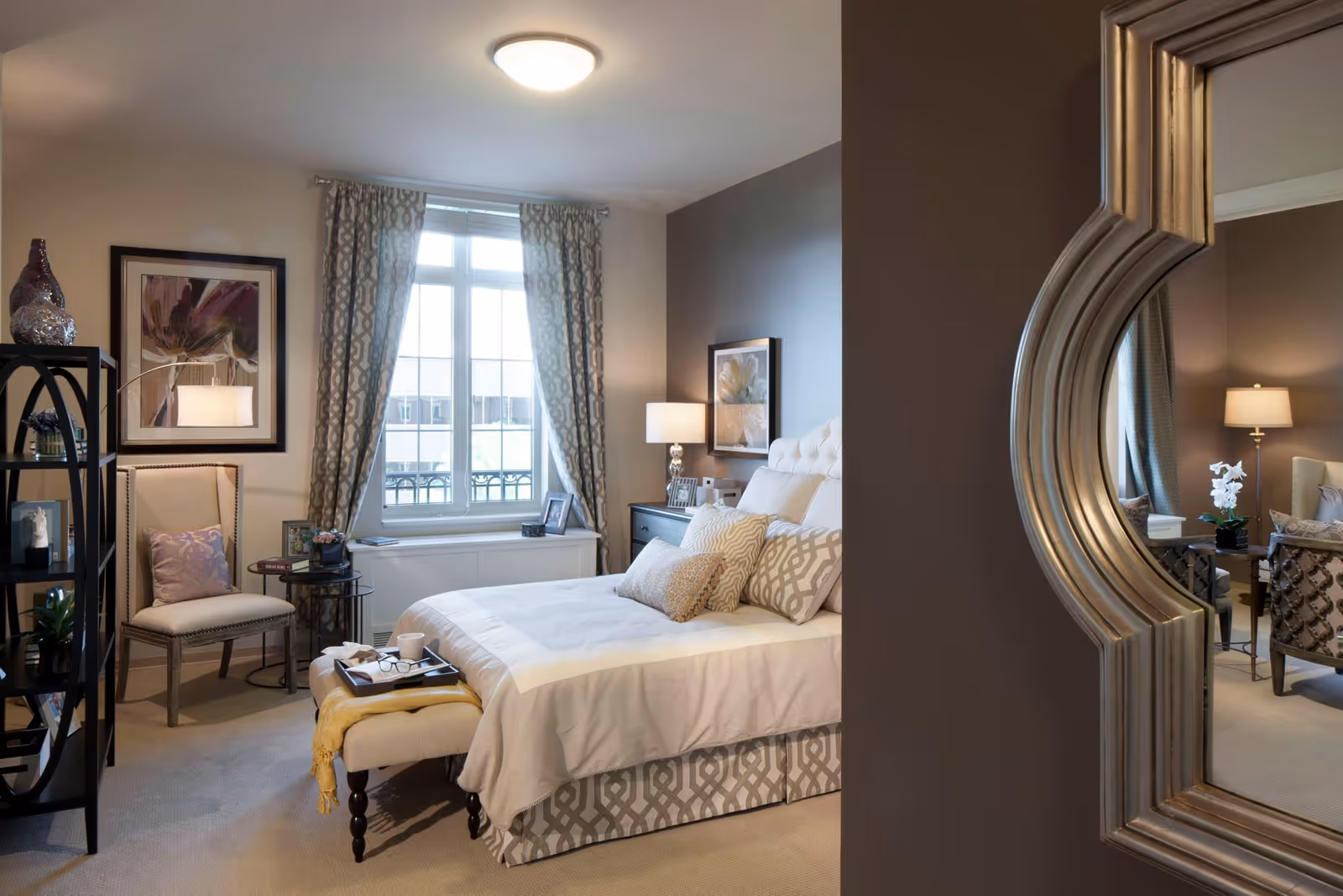 A cozy bedroom in a senior living facility featuring a neatly made bed with multiple decorative pillows, a cushioned bench at the foot of the bed with a tray holding a cup and glasses, a window with patterned curtains letting in natural light, a chair with a pillow, a side table with a lamp, framed artwork on the walls, and a large decorative mirror reflecting part of the room.