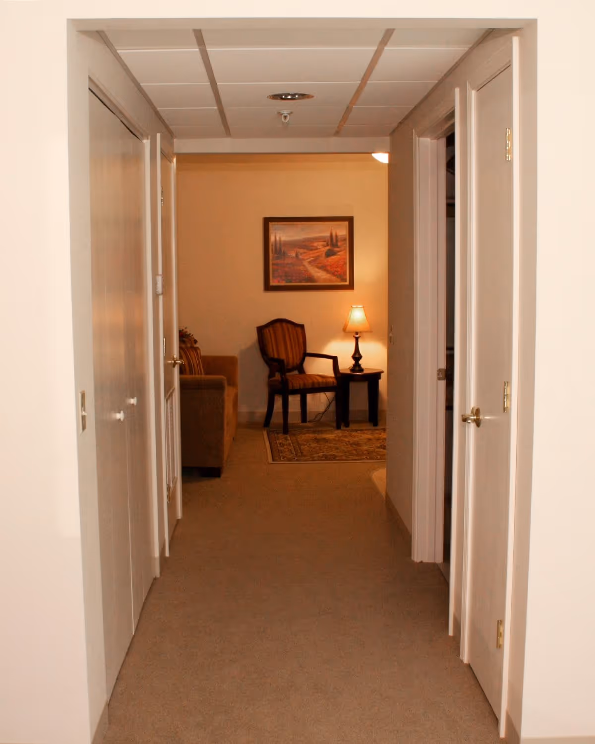 View down a carpeted hallway with white walls and doors on both sides leading to a living area. The living area has a striped armchair, a side table with a lamp, a couch, a framed landscape painting on the wall, and a patterned rug.