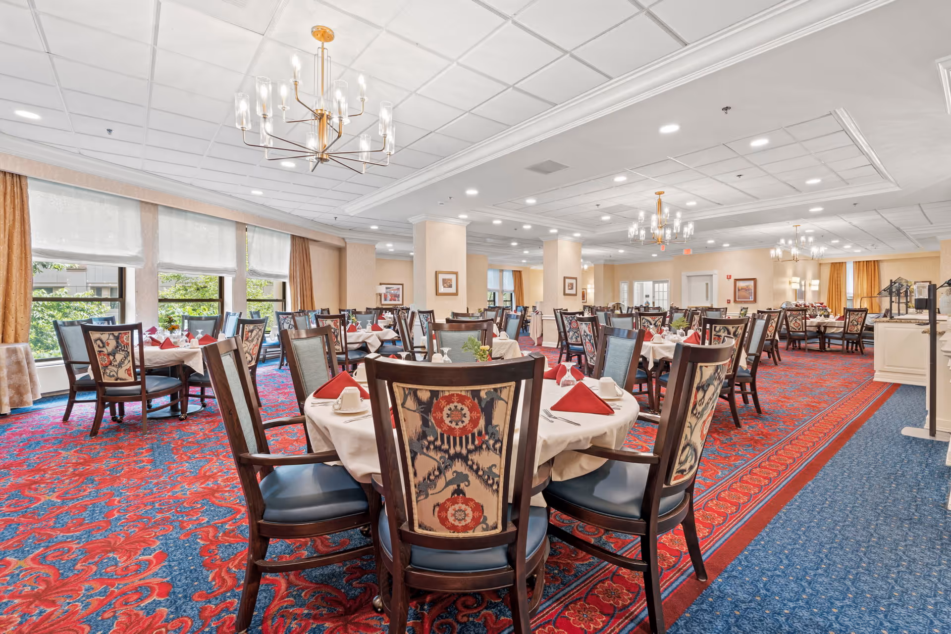 A spacious dining room with multiple round tables covered in white tablecloths, each set with red napkins, white cups, and silverware. The chairs have dark wood frames with patterned upholstery. The room features large windows with beige curtains, red and blue patterned carpet, and elegant chandeliers hanging from a white ceiling with recessed lighting.