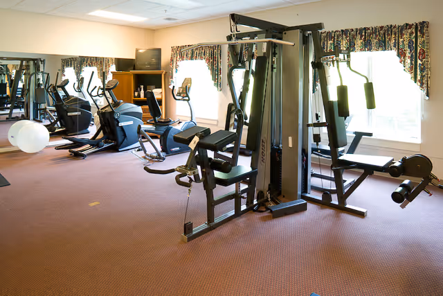 A fitness room with various exercise equipment including stationary bikes, elliptical machines, a multi-function weight machine, and a large exercise ball. The room has carpeted flooring, large windows with floral curtains, and a wall mirror reflecting the equipment.