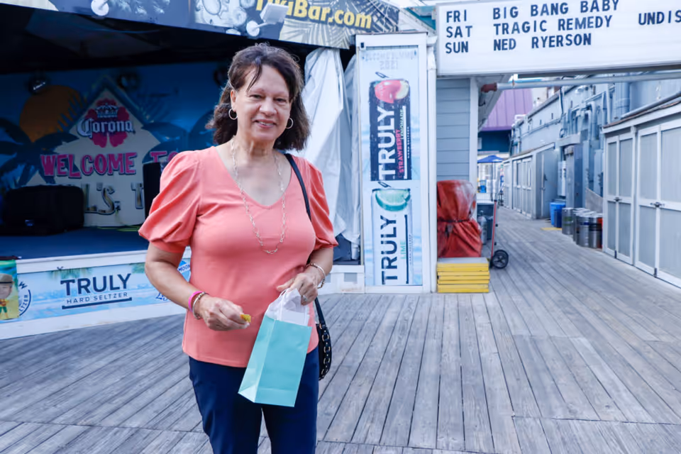 A woman wearing a coral pink blouse and dark pants stands on a wooden boardwalk holding a small light blue gift bag. Behind her is a stage area with a Corona sign and Truly Hard Seltzer advertisements. There is also a marquee sign listing band names and days of the week.