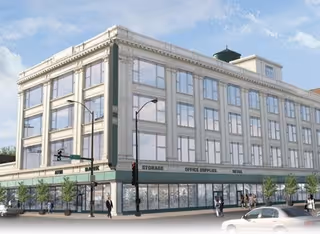 A large, multi-story commercial building with a classic architectural style featuring many windows and decorative cornices. The ground floor has storefronts labeled with signs such as 'STORAGE,' 'OFFICE SERVICES,' and 'RETAIL.' Several pedestrians are walking on the sidewalks around the building, and a car is visible in the foreground. The sky is clear with a few clouds.