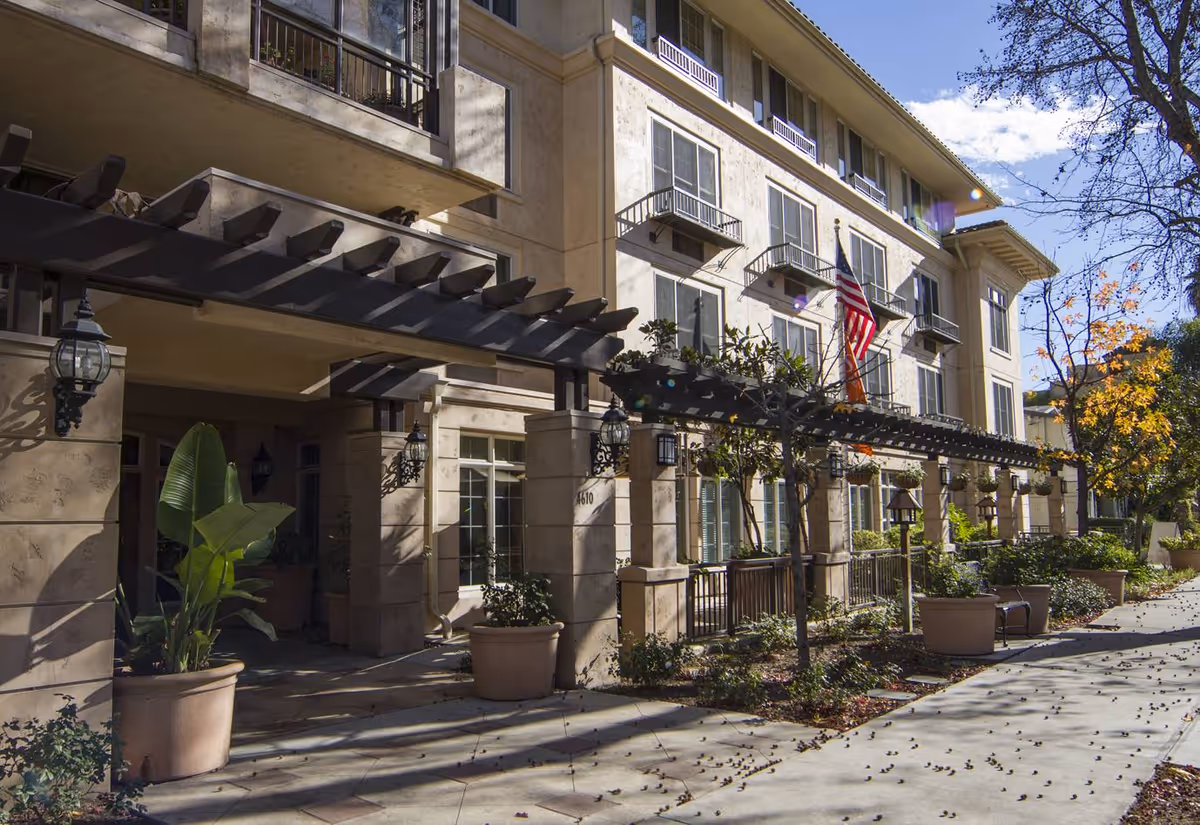 Exterior view of a multi-story senior living facility building with beige walls, multiple windows, and small balconies. There is a pergola structure with hanging plants along the entrance walkway. Potted plants and trees line the sidewalk, and an American flag is displayed near the entrance under a clear blue sky.