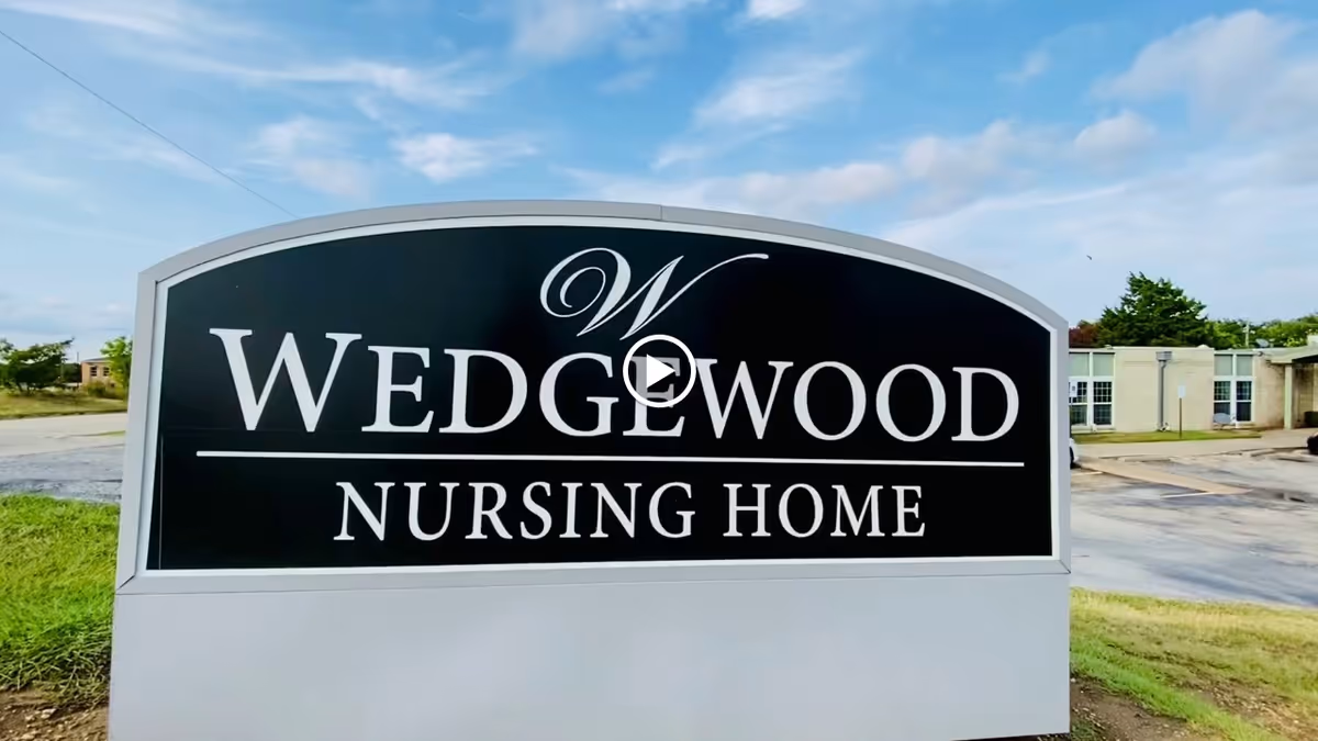 A large black and white sign for Wedgewood Nursing Home situated outdoors with a partly cloudy sky and some buildings in the background.