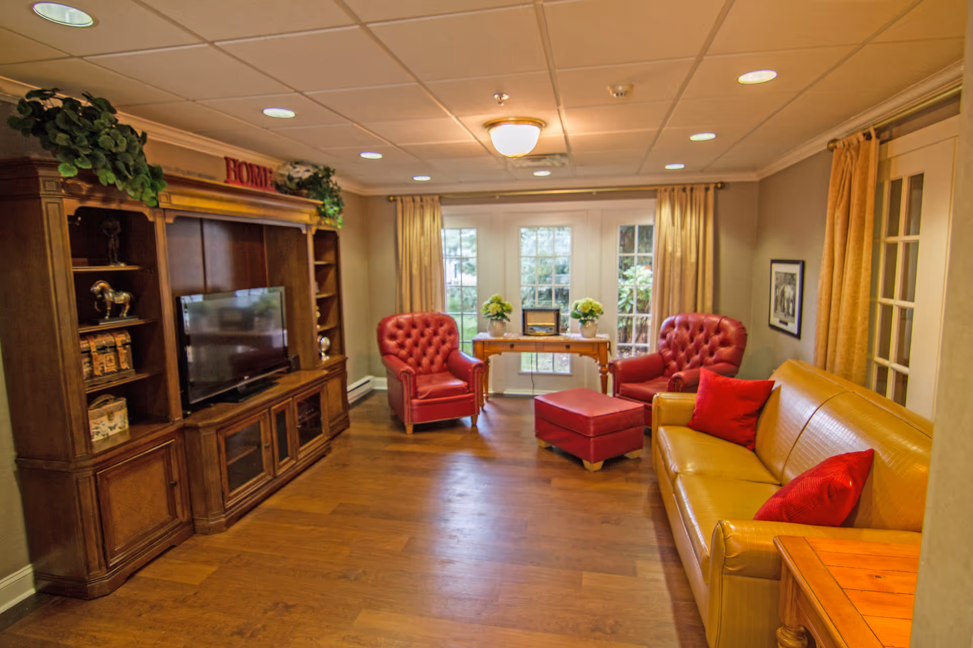 A cozy living room with wooden flooring, featuring a large wooden entertainment center with a TV, two red leather armchairs, a matching red ottoman, a yellow leather sofa with red pillows, a wooden side table, and a wooden console table with potted plants in front of glass doors with beige curtains.