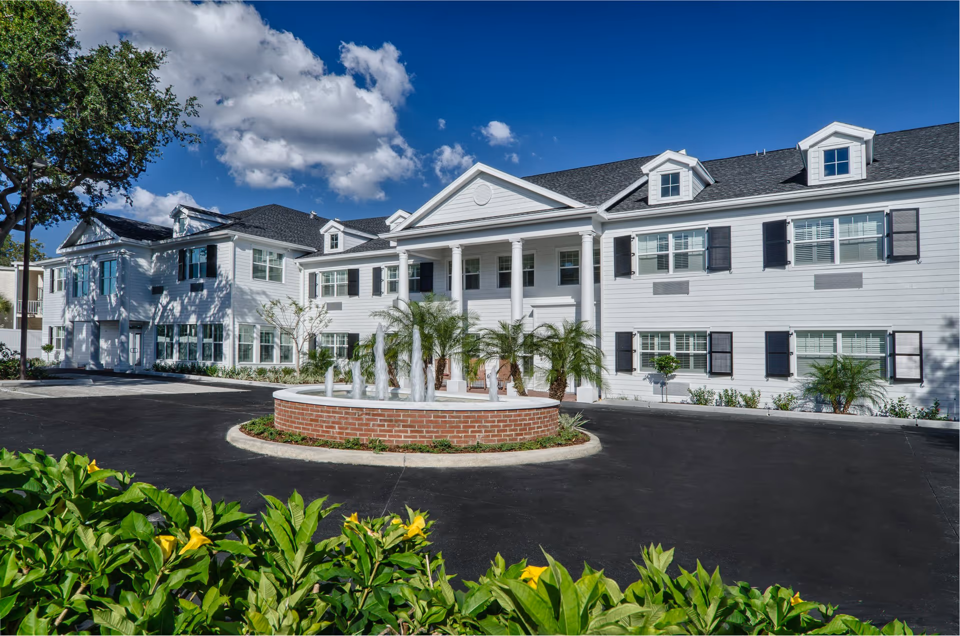 Exterior view of a large two-story white building with black shutters and a dark roof under a blue sky with clouds. The building has a prominent entrance with white columns and a circular brick fountain with water jets in front. Green plants and yellow flowers are visible in the foreground.