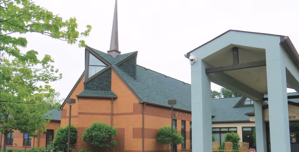 Exterior view of a brick building with a steep green roof and a tall spire, surrounded by trees and shrubs. There is a covered entrance structure with a security camera mounted on it.