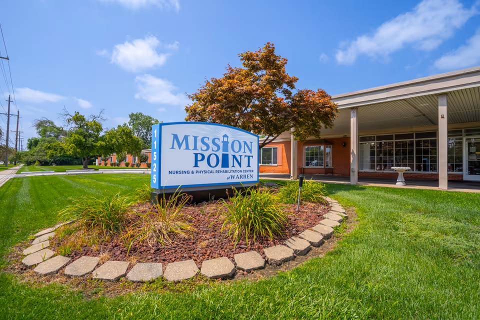 Exterior view of Mission Point Nursing & Physical Rehabilitation Center of Warren with a large sign displaying the facility's name surrounded by landscaping, green grass, and a tree under a blue sky with some clouds.