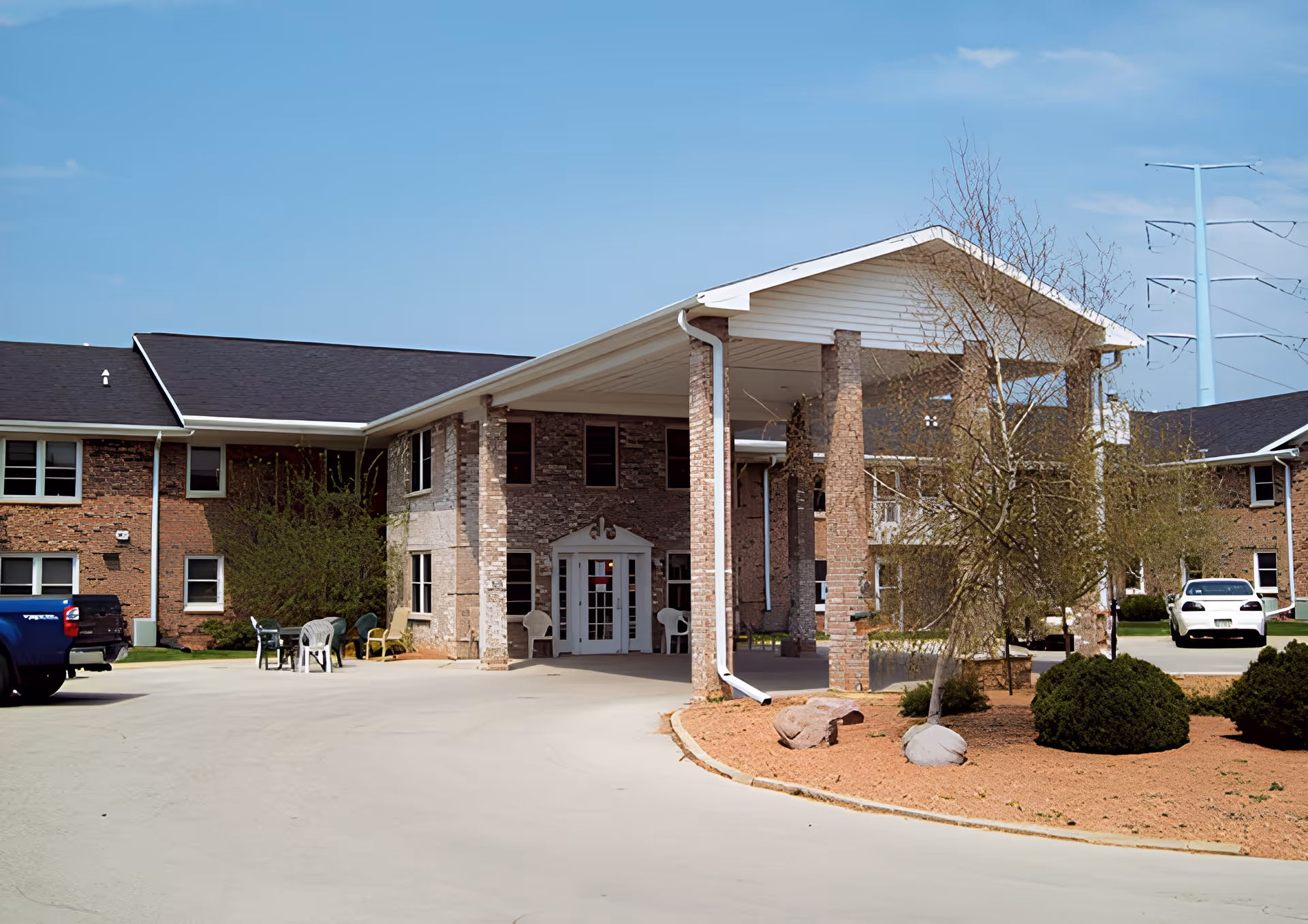 Exterior view of Birch Creek senior living facility showing a brick building with a covered entrance supported by brick columns, a driveway, parked vehicles, and landscaped area with bushes and trees under a clear blue sky.