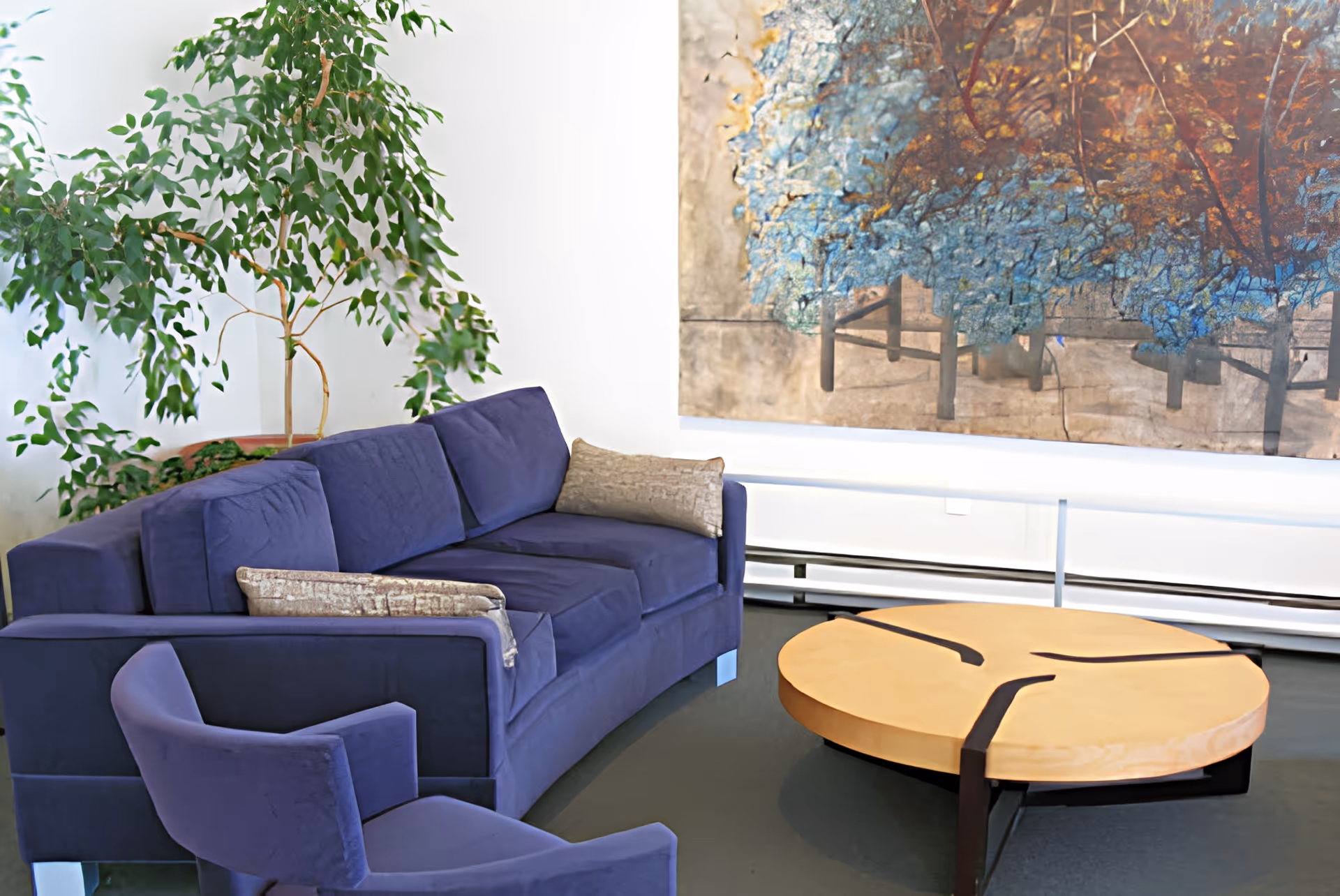 A modern living room with a deep blue sofa and matching armchair, beige cushions, a round wooden coffee table with black metal accents, a large green potted plant, and a colorful abstract painting on the white wall behind the sofa.