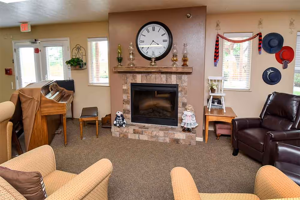 A cozy living room area with a stone fireplace in the center, topped with a large round clock and decorative glass lamps. To the left, there is a covered piano with a bench, and to the right, a dark brown leather recliner next to a wooden side table with a white chair on top. The walls have windows with blinds and decorative hats hanging on the right side. Beige armchairs with patterned pillows are partially visible in the foreground.