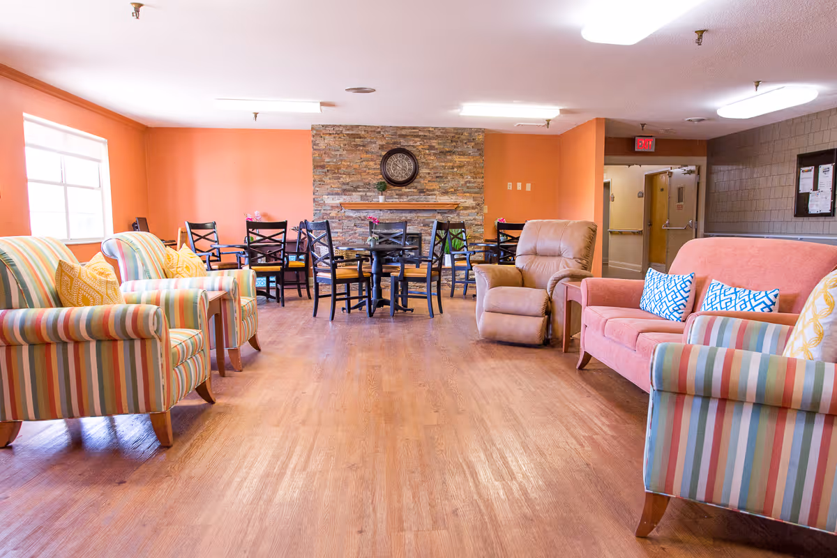 A spacious common area in a senior living facility with wooden flooring, colorful striped armchairs, a pink sofa with patterned cushions, a beige recliner, and several black dining chairs around tables. The back wall features a stone accent with a clock and small plants on a shelf. The room is well-lit with natural light from a window on the left and ceiling lights.