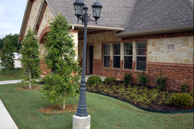 Exterior view of a single-story brick and stone building with a sloped roof, several windows, and a sign that reads 'BLOK E'. In front of the building is a landscaped area with small trees, bushes, and a black lamp post on a concrete base. A sidewalk runs alongside the building and the grass is neatly trimmed.
