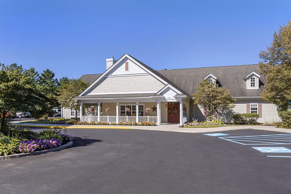 Exterior view of a single-story building with beige siding and a gray shingled roof under a clear blue sky. The building has a covered porch with white railings and hanging flower baskets. There are landscaped flower beds with colorful flowers and trees around the building. A paved driveway and parking area with handicap parking spaces are visible in front.