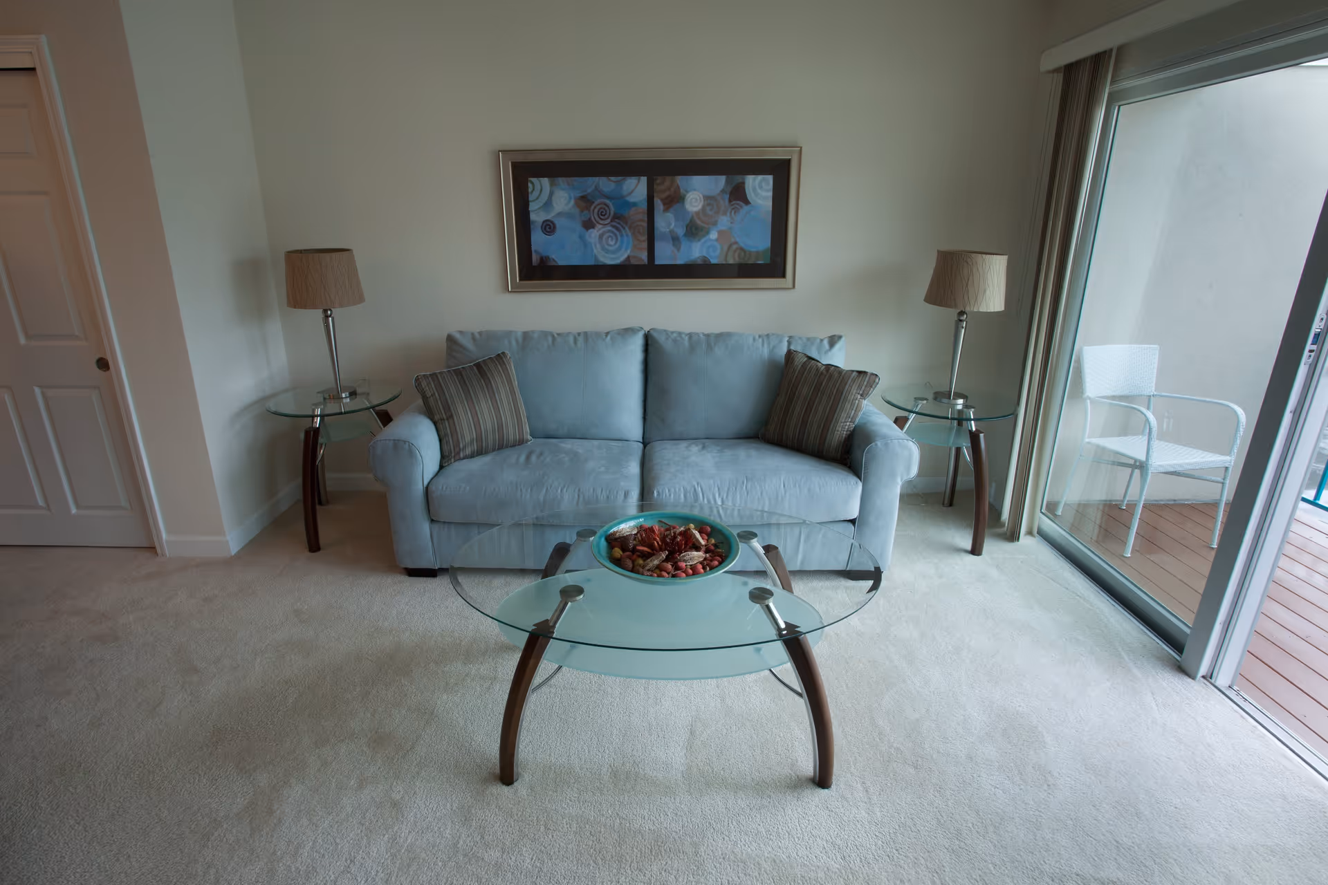 A living room with a light blue sofa adorned with two striped pillows, flanked by two glass side tables each holding a lamp with a beige shade. In front of the sofa is a glass coffee table with a decorative bowl filled with potpourri. A framed abstract artwork hangs on the wall above the sofa. To the right, there is a sliding glass door leading to an outdoor area with a white chair on a wooden deck.
