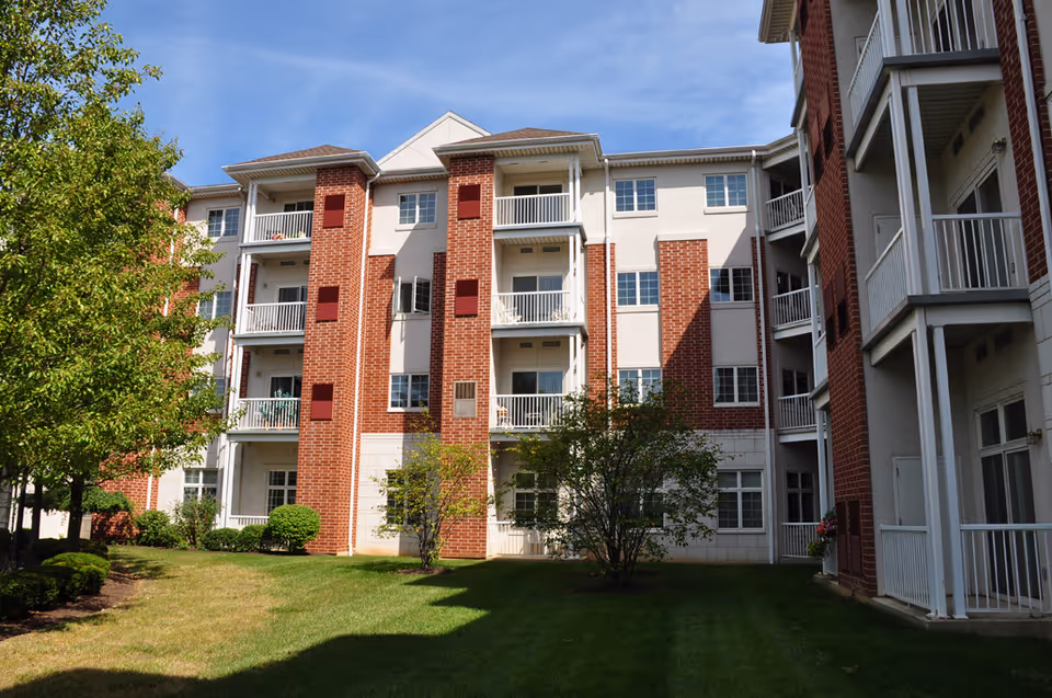 Exterior view of a multi-story senior living building with red brick and white siding, featuring balconies and windows. The building is surrounded by green grass, trees, and shrubs under a clear blue sky.