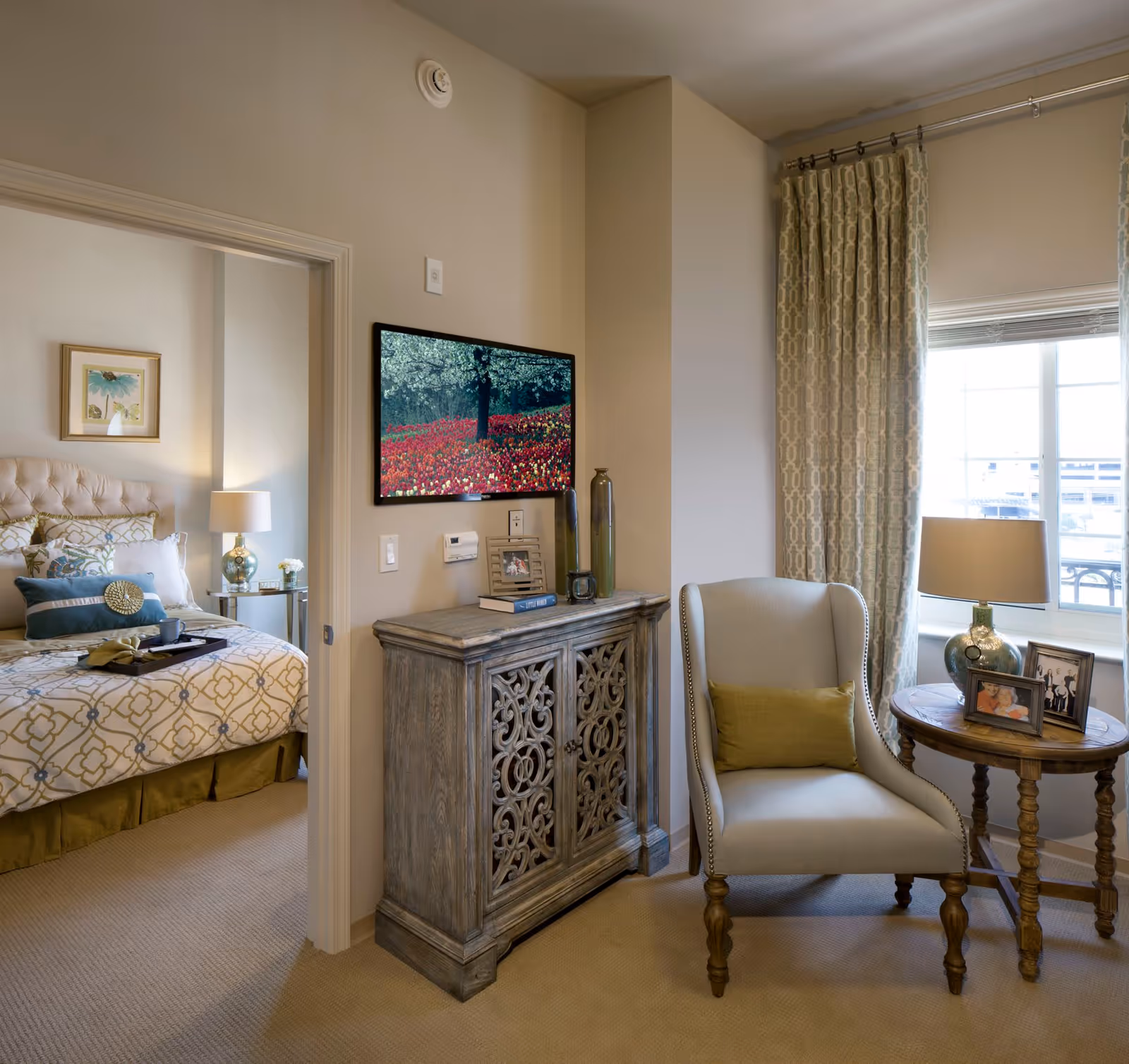 A cozy senior living space featuring a beige armchair with a green cushion next to a round wooden side table with a lamp and framed photos. A decorative wooden cabinet with intricate carvings is against the wall, above which a flat-screen TV displays an image of a tree and a field of red flowers. In the adjoining room, a bed with patterned bedding and multiple pillows is visible, along with a bedside table and lamp. The room has beige walls, carpeted floors, and a window with patterned curtains letting in natural light.