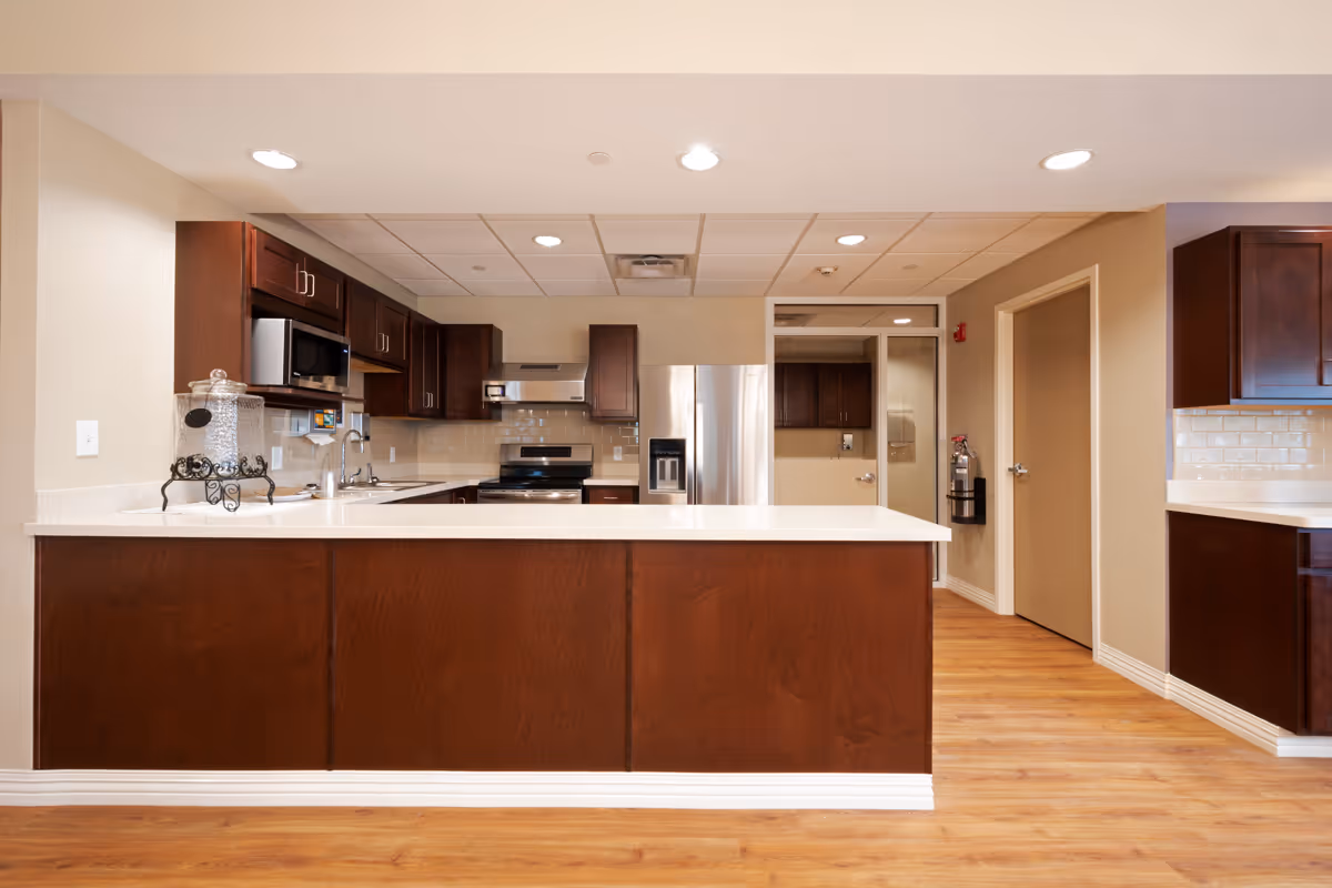 A modern kitchen with dark wood cabinets, a large island with a white countertop, stainless steel appliances including a refrigerator, stove, and microwave, and light wood flooring. The kitchen is well-lit with recessed ceiling lights and has beige walls and a tiled backsplash.