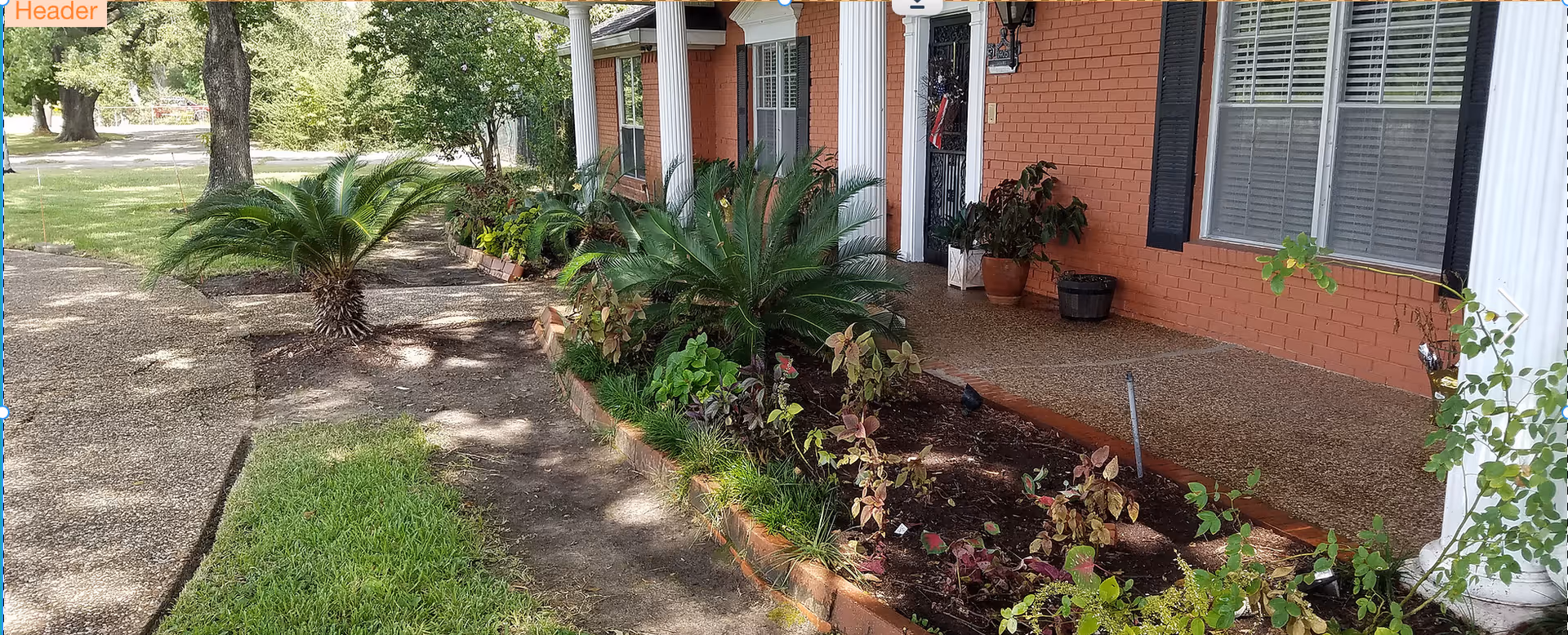 Front porch area of a brick building with white columns, a garden bed with various plants, and a walkway surrounded by grass and trees.