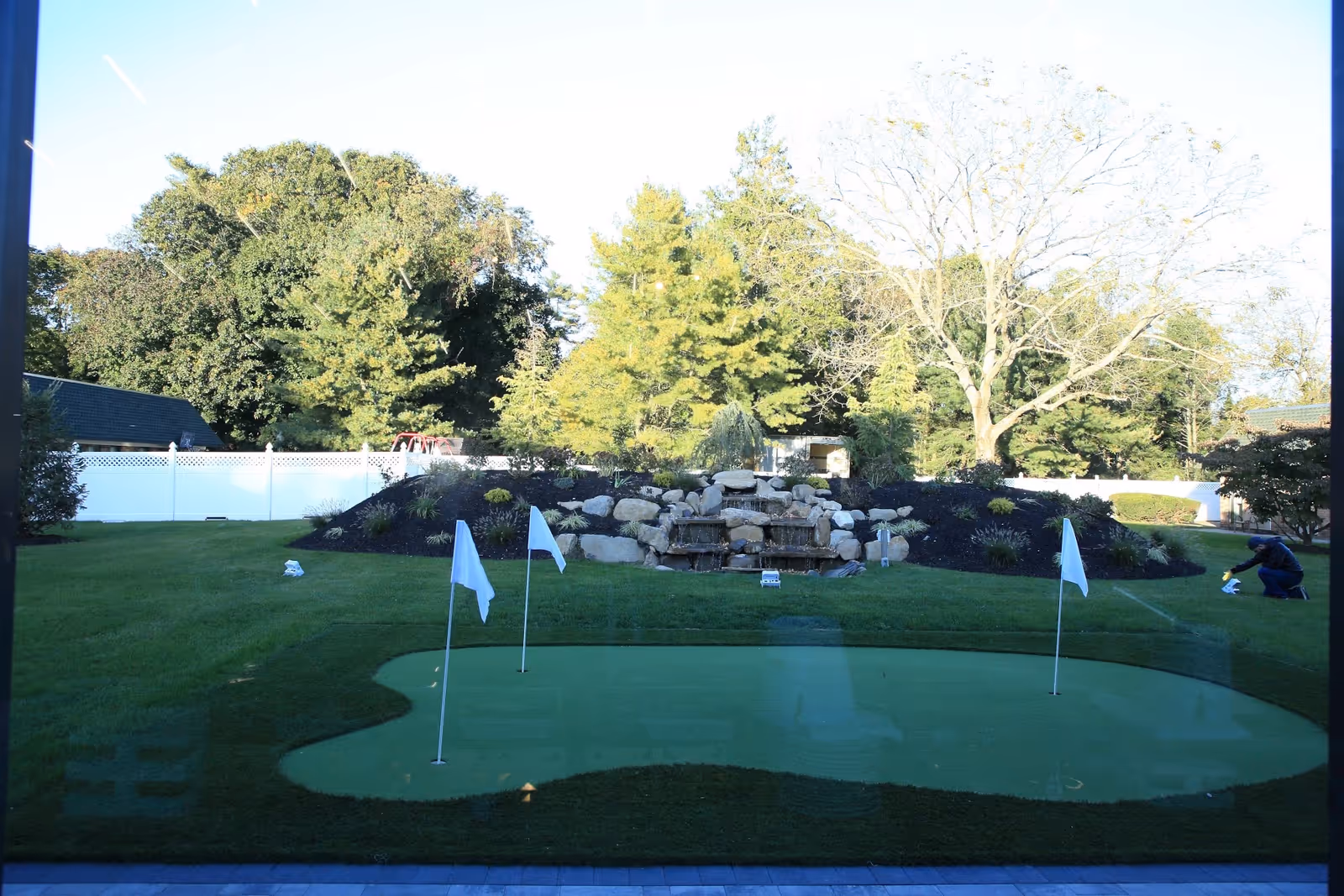 Outdoor putting green with four white flags on poles, surrounded by well-maintained grass and landscaping including a rock waterfall feature and various trees in the background. A person is seen tending to the garden area on the right side.